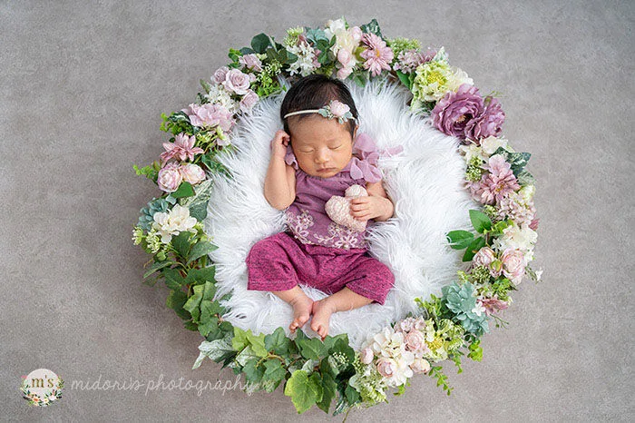 造花のリースの中で眠る新生児。紫のニット衣装と花のヘアアクセサリー、白いファーを合わせたニューボーンフォト