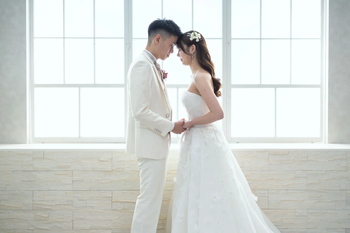 窓の前で向かい合わせになり、頭を合わせる新郎新婦。白い衣装を着ている。