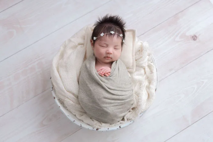 ベージュのおくるみに巻かれ、白い器の中で眠る新生児。小花のヘアアクセサリーを合わせて真上から撮影