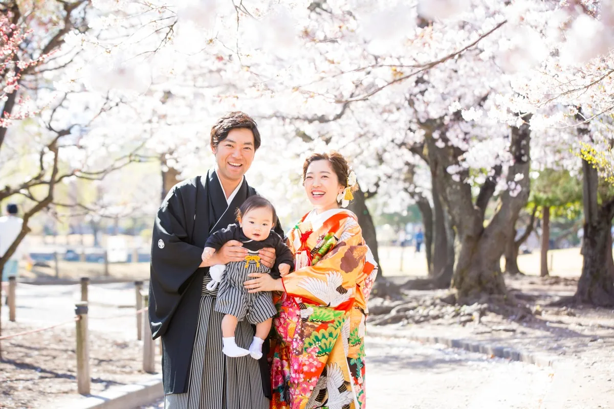 桜の下で、赤ちゃんを抱いた紋付袴の新郎と色打掛の新婦。3人で正面を向いて笑っている