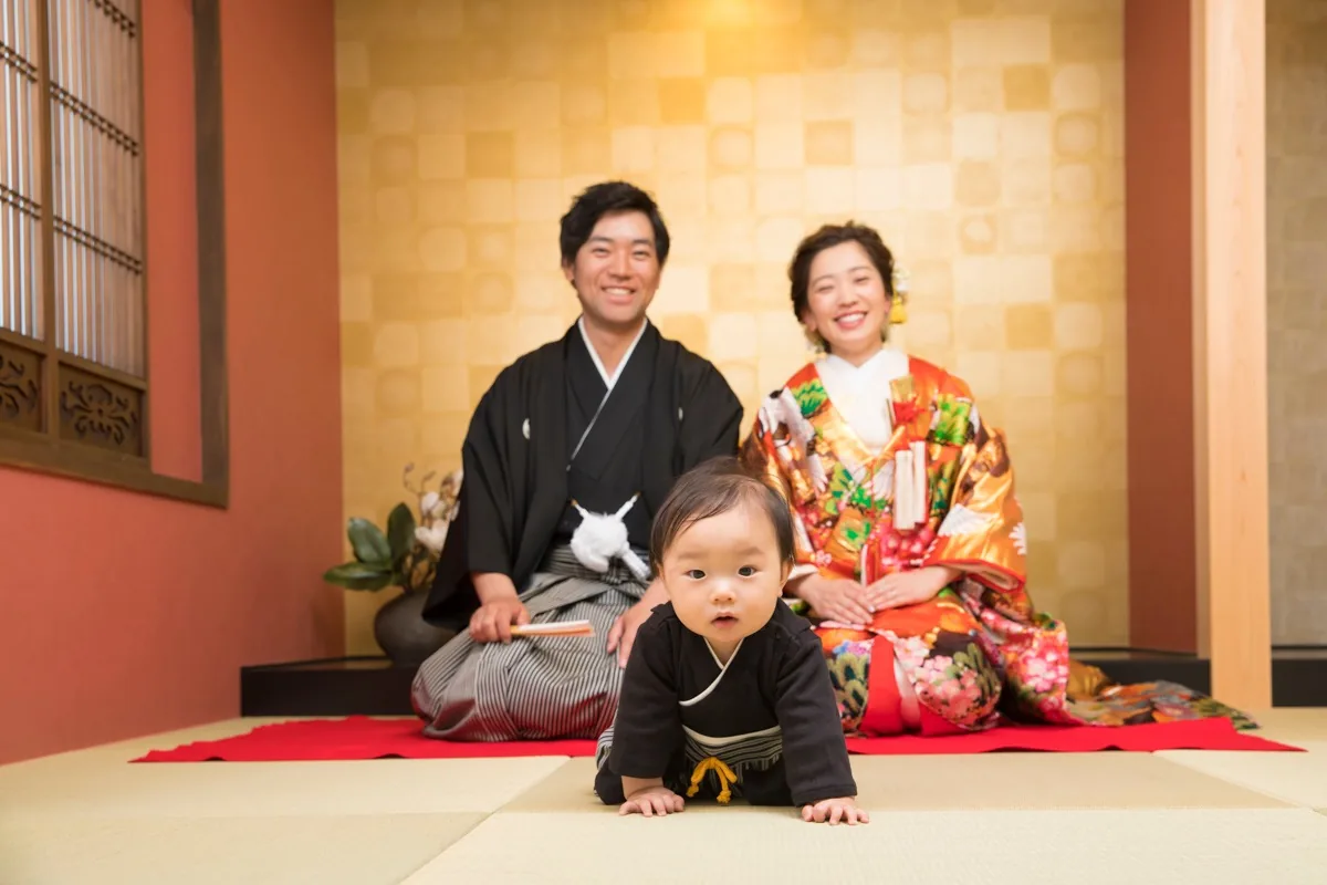 和室の畳の上をハイハイする赤ちゃんと、後ろに座る紋付袴の新郎と色打掛の新婦