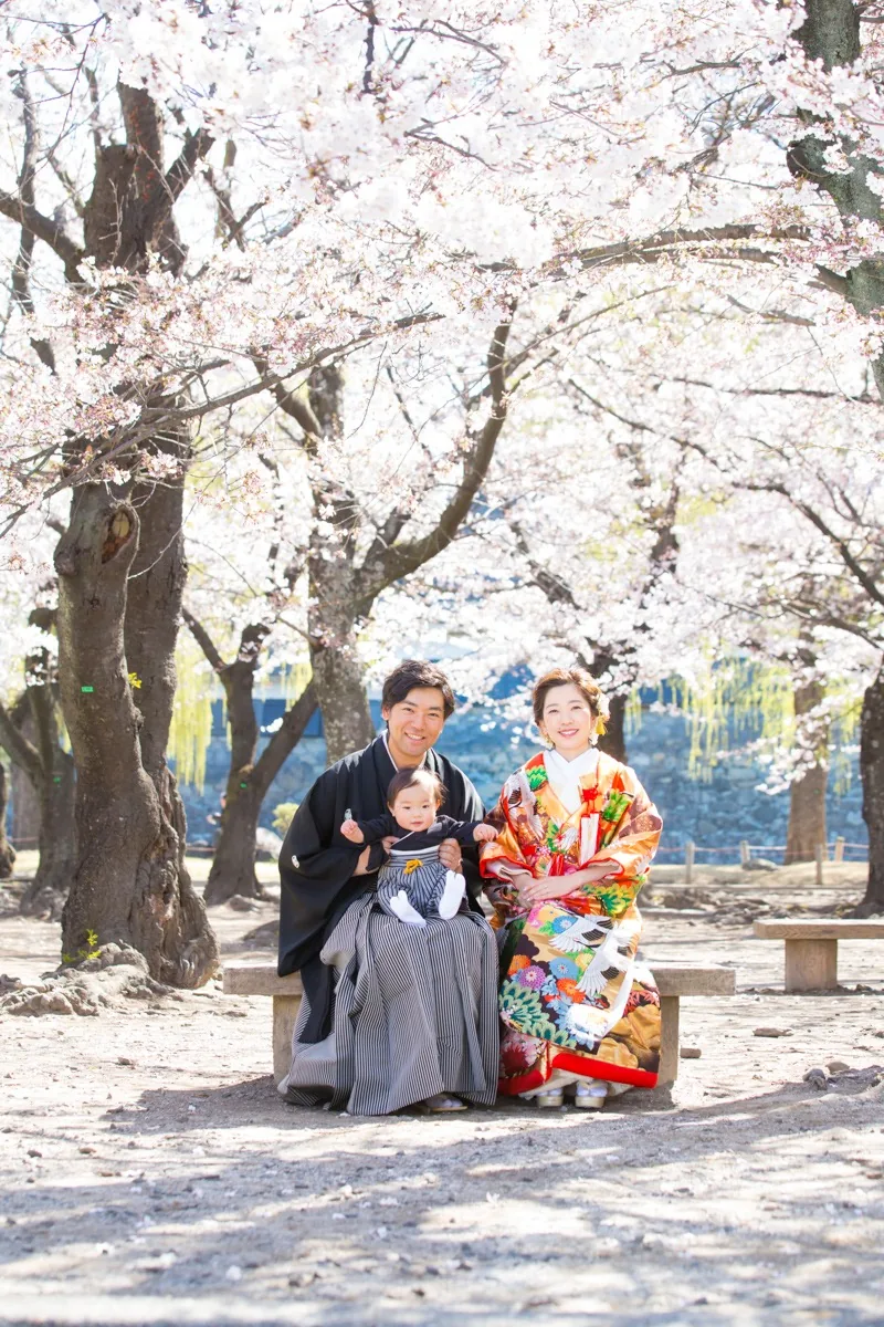 松本城の石垣を背に、桜の木の下のベンチに座る家族3人。新郎が赤ちゃんを膝に乗せている