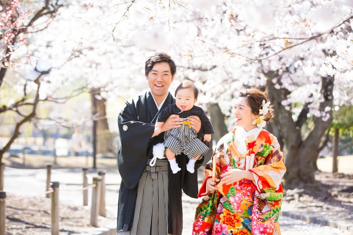 桜の下で、新郎が赤ちゃんを抱き上げ、隣で色打掛の新婦が笑っている