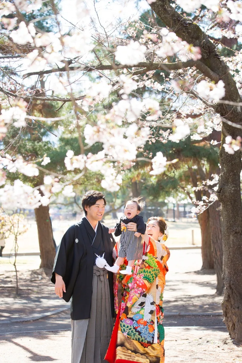 桜の木の下で、新郎新婦が赤ちゃんを高く抱き上げて、赤ちゃんが声を出して笑っている