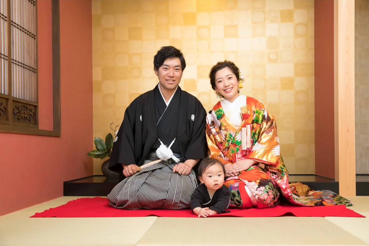 和室で正座する紋付袴の新郎と色打掛の新婦。手前で赤ちゃんが畳に腹ばいになっている