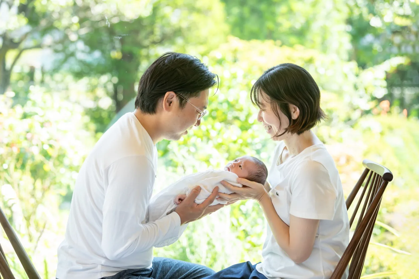 庭の緑を背に、おくるみに包んだ赤ちゃんを両親で見つめる。松本のライフスタイルニューボーンフォト