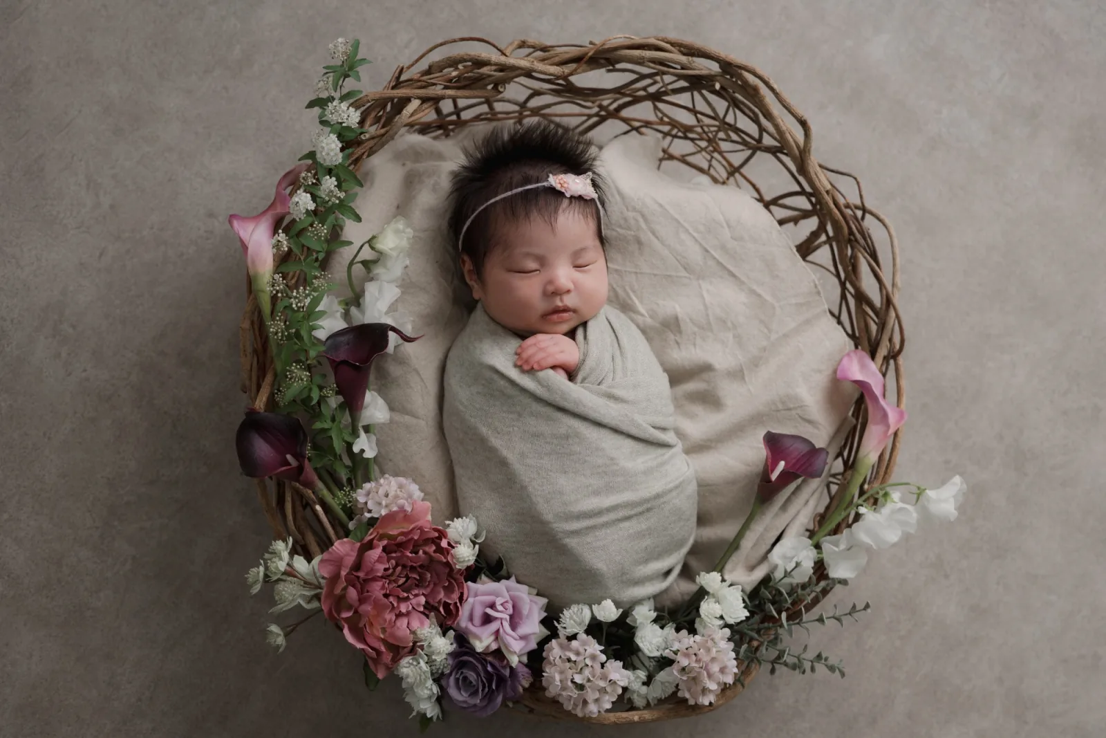 細い枝で編んだかごの中で、グレーベージュのおくるみに包まれて眠る新生児。ピンクの花のヘアバンドをつけ、まわりにカラーやバラの花を添えて真上から