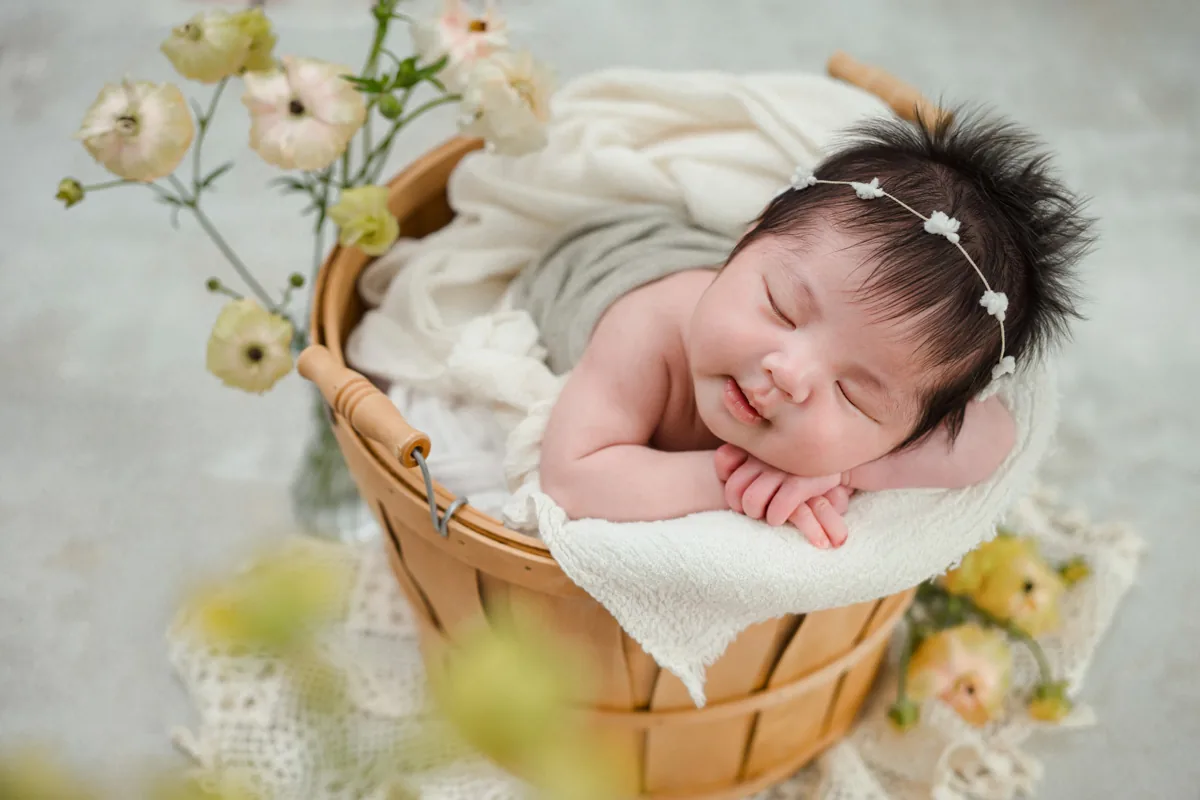 木のバスケットの中で、白いブランケットにほほをのせて、ほほえむように眠る新生児。白い小花のヘアバンドをつけ、淡い黄色とピンクの花を添えて